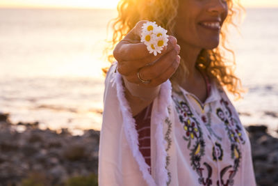 Midsection of woman holding white flowers while standing at beach during sunset