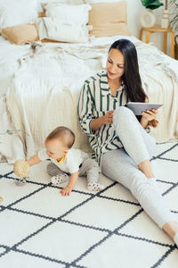 A pretty mom with a computer tablet sits on the floor and spends time playing with a toddler baby. 