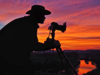 Silhouette man photographing on field against orange sky