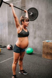 Side view of pregnant female athlete lifting barbell during training in modern gym and looking away