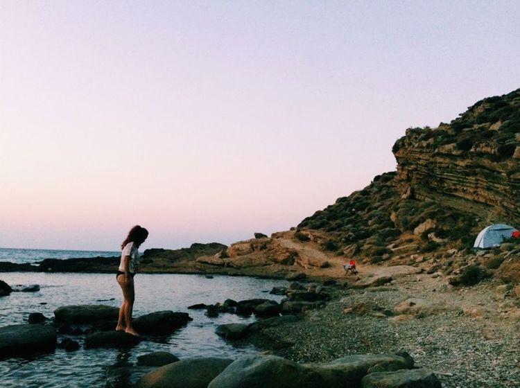 MAN STANDING ON ROCK BY SEA AGAINST SKY