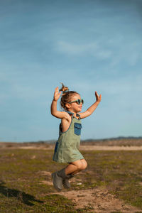 Side view of content little girl in overalls and sunglasses jumping with outstretched arms above meadow and enjoying summer on sunny day in countryside