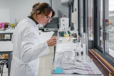 Side view of mature female scientist with clipboard examining glassware while working in modern chemistry laboratory