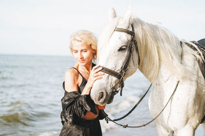 Young blonde woman in black clothes riding white horse on seascape background