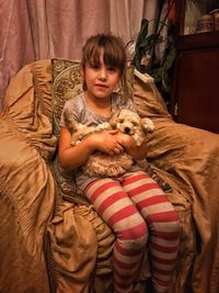 Portrait of girl holding puppy while sitting on armchair