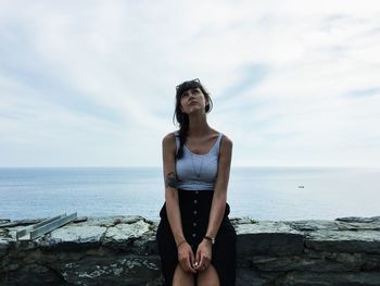 Young woman sitting by beach against sky
