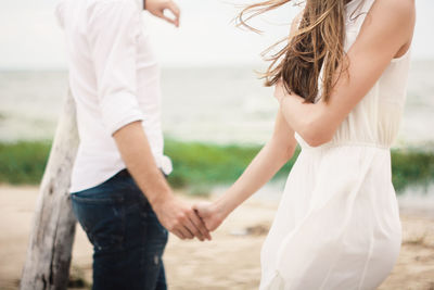 Midsection of couple holding hands while standing by sea