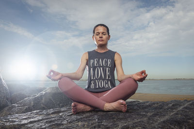 Woman sitting on rocks by the sea wearing a t shirt with 'peace love and yoga' slogan on it.