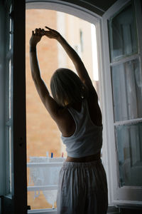 Back view of anonymous female in pajamas standing near window and stretching after awakening at home while enjoying morning