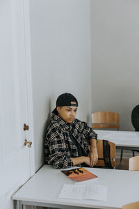 Thoughtful young man sitting at desk while leaning on white wall in classroom