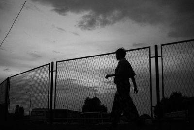 Silhouette man standing by fence against sky during sunset