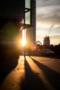 Silhouette city street by buildings against sky during sunset