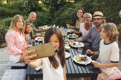 Happy family and friends looking while girl taking selfie from mobile phone at back yard during garden party