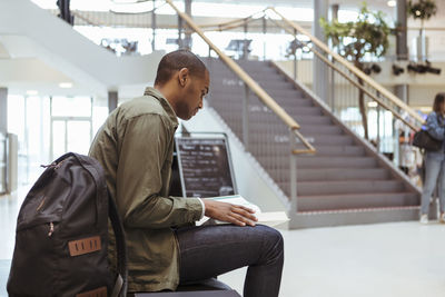 Young student reading book while sitting in corridor at high school