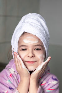 A girl in the bathroom with a towel on her head washes her face with cleansing foam with her hands