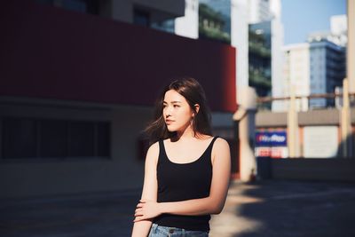 Young woman looking away while standing on road against buildings in city