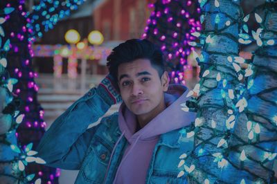 Portrait of young man standing by illuminated christmas tree at night