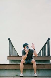 Man photographing with camera on steps while waving against clear sky