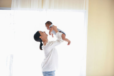 Side view of mother picking up son while standing against window at home