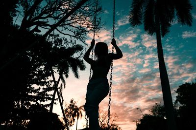 Low angle view of silhouette woman standing on swing against sky during sunset