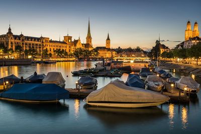 Boats moored in river by illuminated city against clear sky