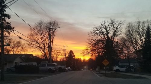 Road by bare trees against sky during sunset