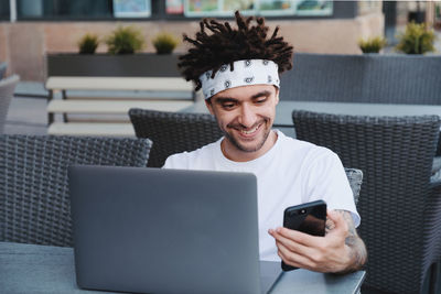 Smiling young man using phone and laptop while sitting at table