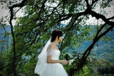 Side view of beautiful bride holding bouquet while standing against trees