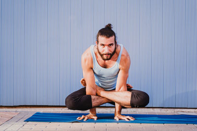 Full length of man performing yoga on exercising mat against wall