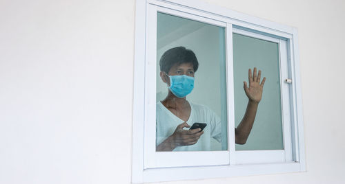 Close-up of mature man wearing flu mask holding phone seen through window