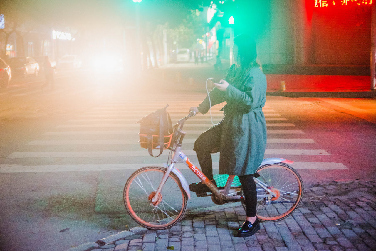 bicycle, real people, transportation, land vehicle, mode of transport, street, lifestyles, one person, cycling, full length, outdoors, city life, riding, rear view, night, city, road, leisure activity, women, built structure, architecture, illuminated, building exterior, men, warm clothing, people