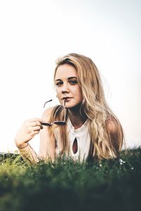 Portrait of young woman holding sunglasses while lying on field against clear sky