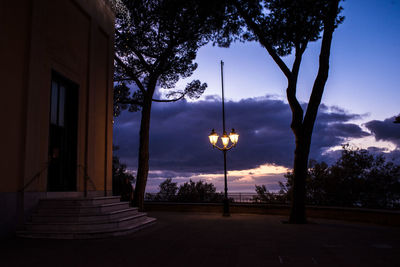 Street light and silhouette trees by building against sky at sunset