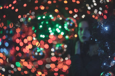 Portrait of young woman standing amidst illuminated christmas tree at night