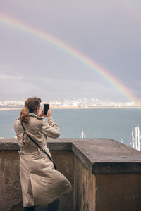 Rear view of woman photographing rainbow over sea against sky