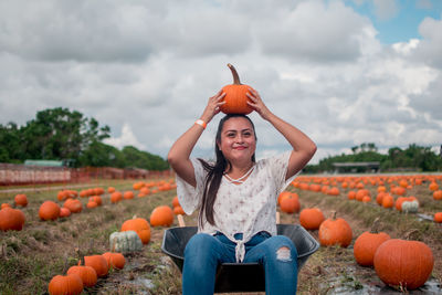 Smiling young woman balancing pumpkin on head while sitting in wheelbarrow at field against sky