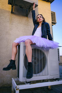 A ballerina in tutu, jacket and boots climbed on the air conditioner and poses against the sky