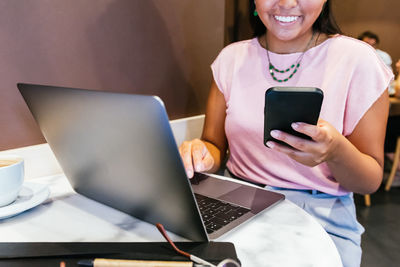 Happy young ethnic self employed lady with long dark hair in casual clothes smiling while messaging on mobile phone sitting at table with laptop during remote work in cafe