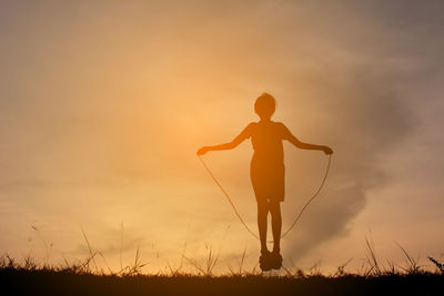 Silhouette girl playing skipping on field against sky during sunset