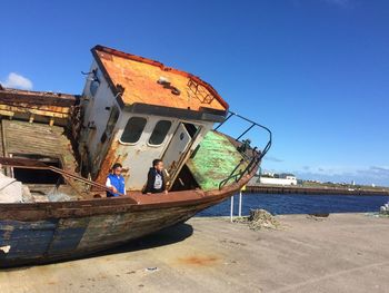 Boys in abandoned boat on pier against blue sky