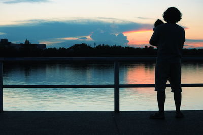 Rear view of silhouette man standing by lake against sky during sunset