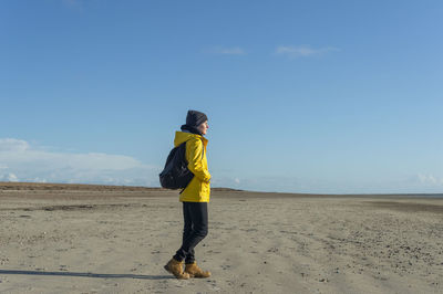 Woman wearing a yellow coat and backpack walking along a beach, sunny autumn day.