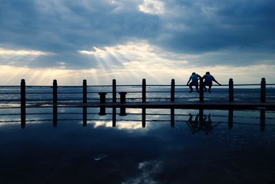 Silhouette men sitting on pier at beach against sky during sunset