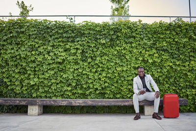 Businessman sitting on a bench with a red suitcase and mobile phone in hand.