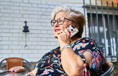 Smiling senior woman in glasses and casual clothes looking away while talking on cellphone near green plants and flowers in garden in summer day