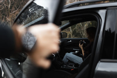 Crop anonymous person opening door of car with woman sitting inside and checking pictures on photo camera during road trip through autumn woodland