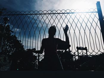 Rear view of silhouette man standing by fence against sky
