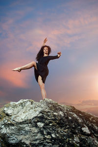 Full length of young woman standing on rock against sky during sunset