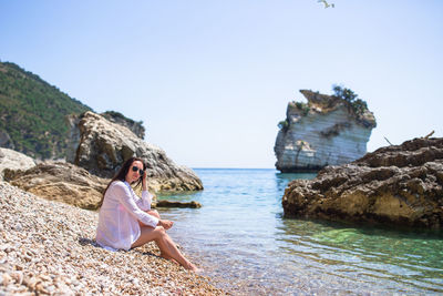 Young woman sitting on rock by sea against clear sky