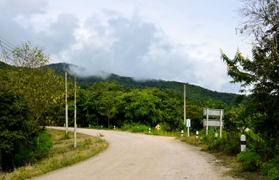 Street amidst trees against cloudy sky